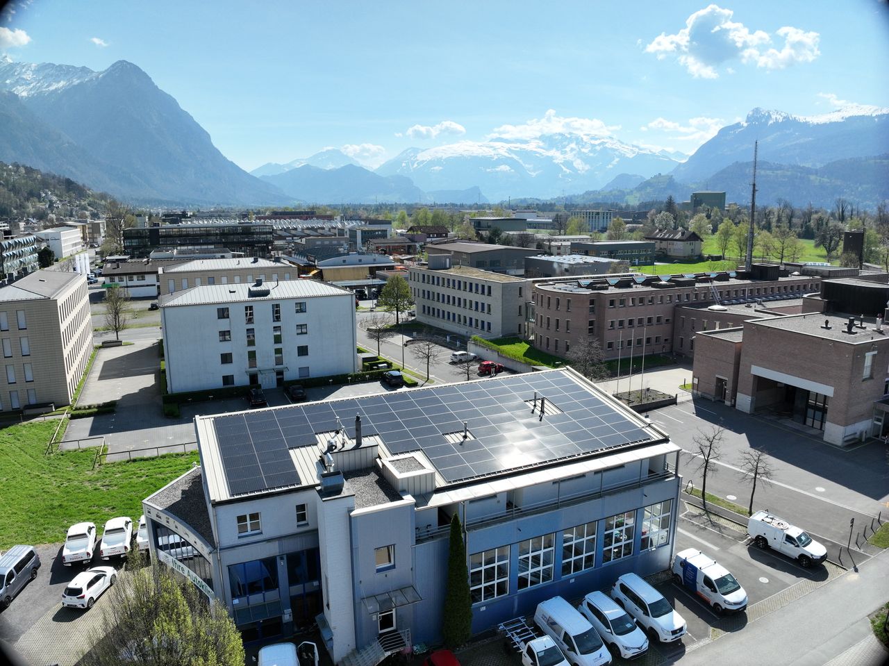 Luftaufnahme eines Gewerbegebäudes, dessen Dach vollflächig mit Photovoltaikmodulen belegt ist, im Hintergrund verschneite Berge