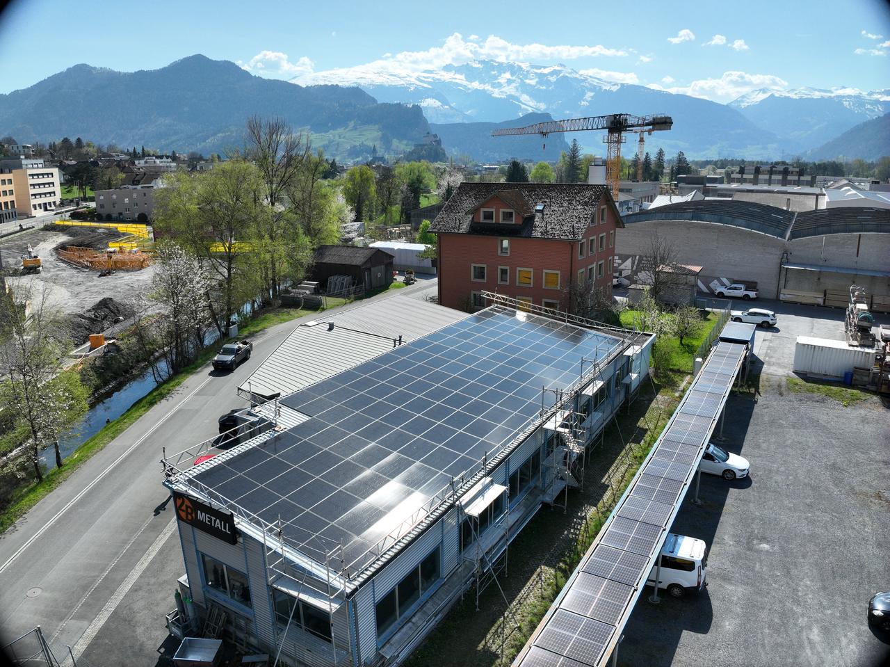 Luftaufnahme einer Gewerbehalle mit vollflächig belegtem Dach und eingerüstetem Baustellenbereich, daneben eine lange Carport-Anlage mit Modulen