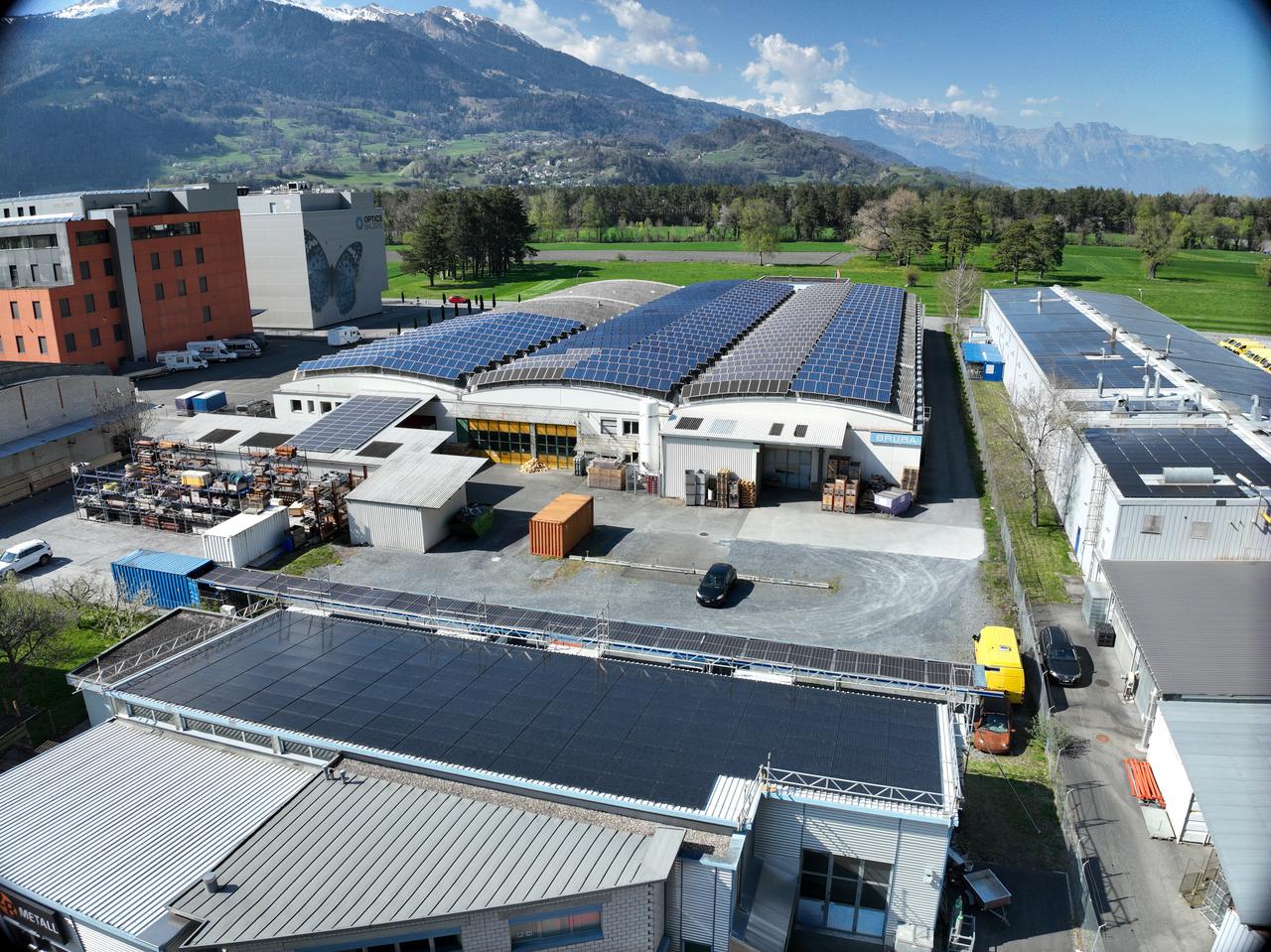 Luftaufnahme eines Industrieareals mit Photovoltaikmodulen auf einem Sheddach und auf mehreren angrenzenden Flachdächern, dahinter Berge